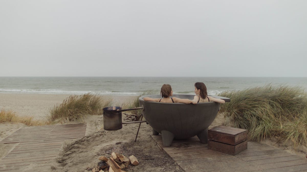 Two women enjoy a serene moment in a round bathtub by the ocean's edge.